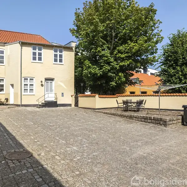 Rækkehus med cremefarvet facade står stille i en brolagt gård. I baggrunden er der store grønne træer og en mur. En simpel, sort udendørs bænk og bord ses nær en affaldscontainer.