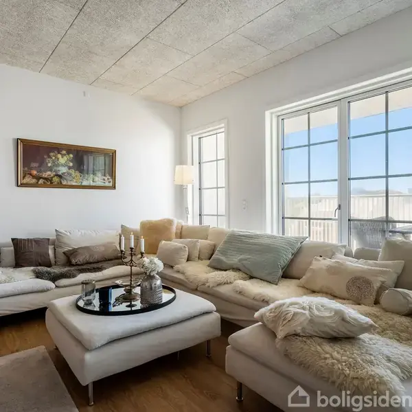 En stor, lys stue med hvide vægge indeholder en stor, beige sofa med bløde puder og tæpper. Et sofabord er i midten, omgivet af glasdøre, der lukker lys ind.