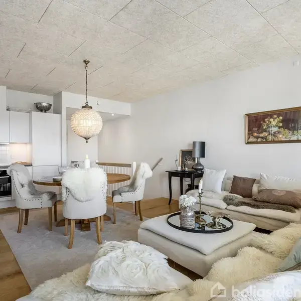 Et hyggeligt opholdsrum med en hjørnesofa fyldt med puder, et sofabord med blomster, en spisebord med stole dækket af pels, samt et åbent køkken i baggrunden. Væggen prydes af et maleri.