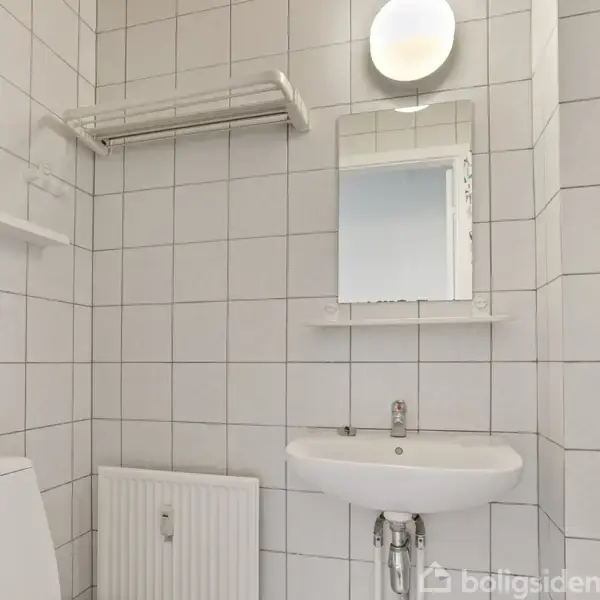 Et badeværelse med hvidt flisevæg, vask med spejl ovenover. En radiator under spejlet og håndklædestang til venstre. Minimalistisk indretning.