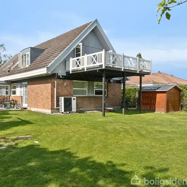 Toetagers hus står stille i solrigt haveområde med velholdt græsplæne og omgivet af træer. En overetage har en balkon understøttet af søjler, ved siden af en rødbrun skur.