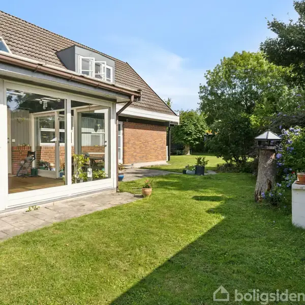 Et hus med en stor glasveranda åbner ud til en velplejet græsplæne med frodige planter og buske. En stenterrasse er foran huset. Sollys belyser området.