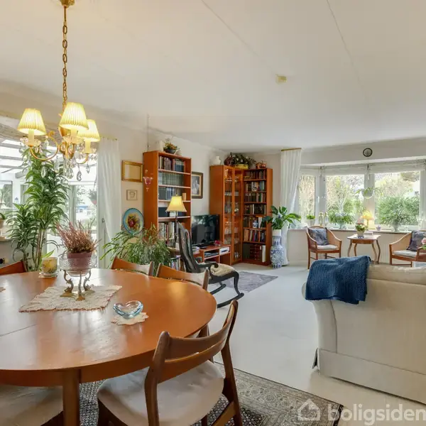 Rundt træbord med stole står i en lys stue; omkring er sofaer, reoler og planter. Store vinduer slipper dagslys ind, hvilket skaber en hyggelig atmosfære.