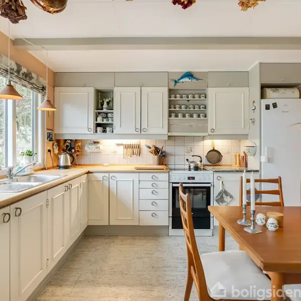 Et køkken med hvide skabe og bordplade i træ. Bordet har lysestager og krus. Vinduerne har planter, og der hænger tørrede blomster fra loftet. Køleskab til højre.