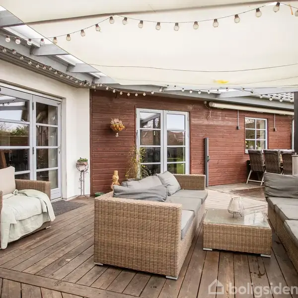 Udemøbler står på en terrasse af træ med sofaer og borde. En markise er spændt over terrassen, og lyskæder hænger i luften. Terrasse omgivet af trævægge og store vinduer.
