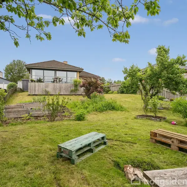 Hus står stille i baggrunden med terrasse og buske omkring. Træpaller fungerer som siddepladser i forgrunden, omkranset af en grøn græsplæne og træer.