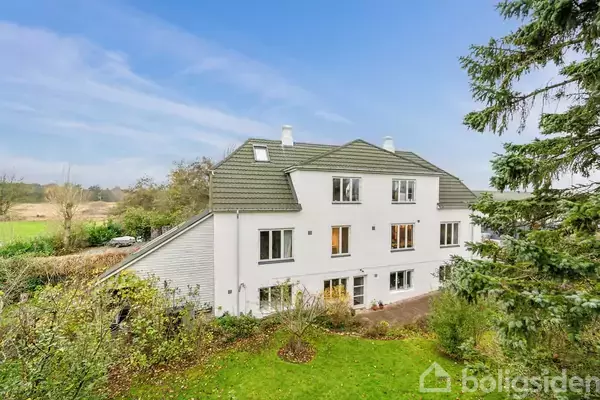 Hvid fleretagers bygning omgivet af grønne træer og buske på en græsplæne; moderne arkitektur med flere vinduer, beliggende i landligt område med blå himmel i baggrunden.