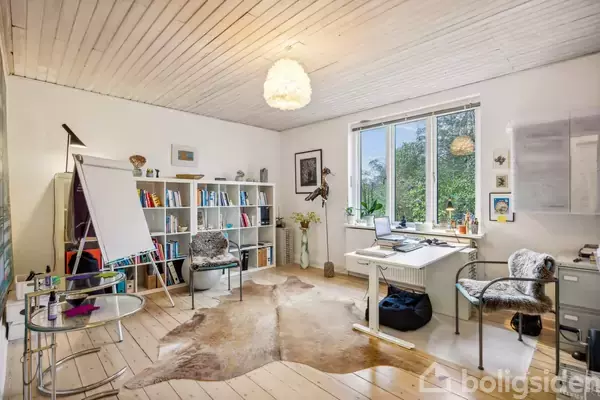 Et lyst kontor med et skrivebord og bærbar computer ved et vindue. En reol med bøger og en flipover står ved siden af. Et tæppe ligger på trægulvet.