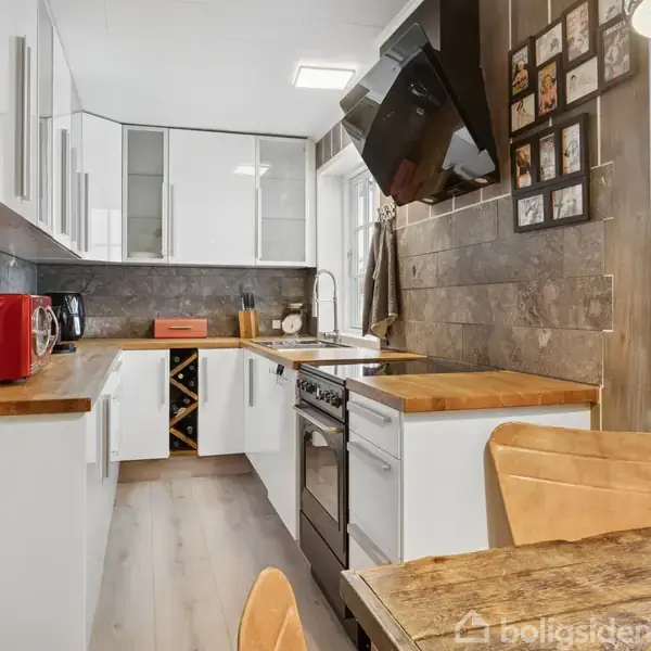 Et moderne køkken har hvide skabe, træborde og apparater som rød mikrobølgeovn. En ovn er placeret centralt, omgivet af billeder på væggen. Et vindue viser udsigt til naturen.