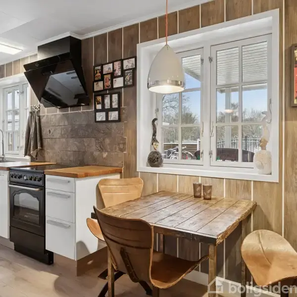 Køkken med træbeklædning. Bord og stole står ved vinduet. Komfur og emhætte er mod væggen. Indretning: lyst, skandinavisk. Batman-plakat på væggen. Vinduer viser udendørs landskab.