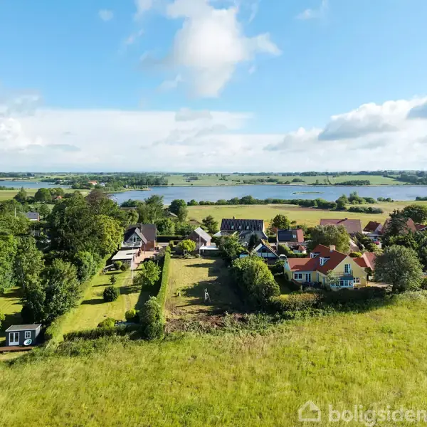 Flere huse ligger i en grøn, landlig omgivelser nær en sø. Træer og marker omringer huse, der har farverige tage. Det er en solrig dag med få skyer på himlen.