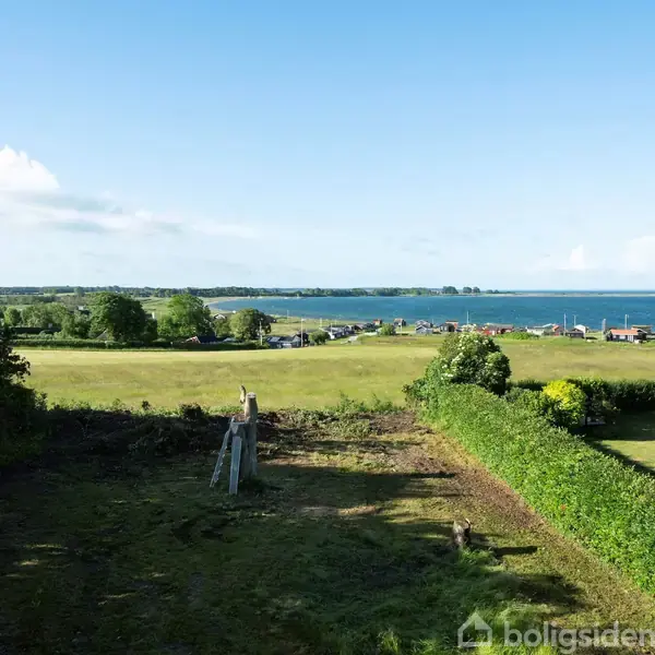 Grøn græsplæne omgivet af tætte hække strækker sig mod en fjord i baggrunden. Små både ses fortøjet tæt ved strandkanten. Over himlen er der spredte, hvide skyer.