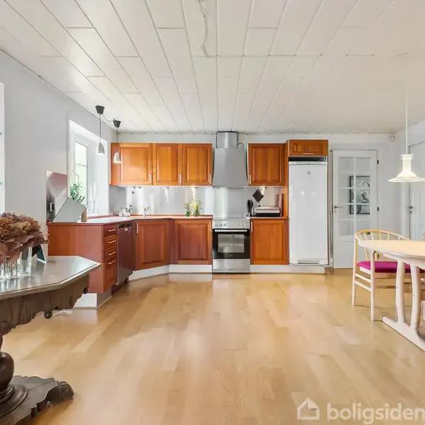 Køkken-alrum med træskabe og moderne apparater, inkluderer et spisebord med stole. Lysekroner hænger fra loftet, og et bord med dekorative genstande står til venstre i et lyst rum.