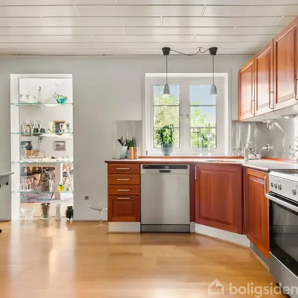 Køkken med træskabe og moderne apparater, herunder en ovn og opvaskemaskine. Rummet har lyst trægulv og et vindue med udsigt til grønne omgivelser. Anretning med vaser ved vinduet.