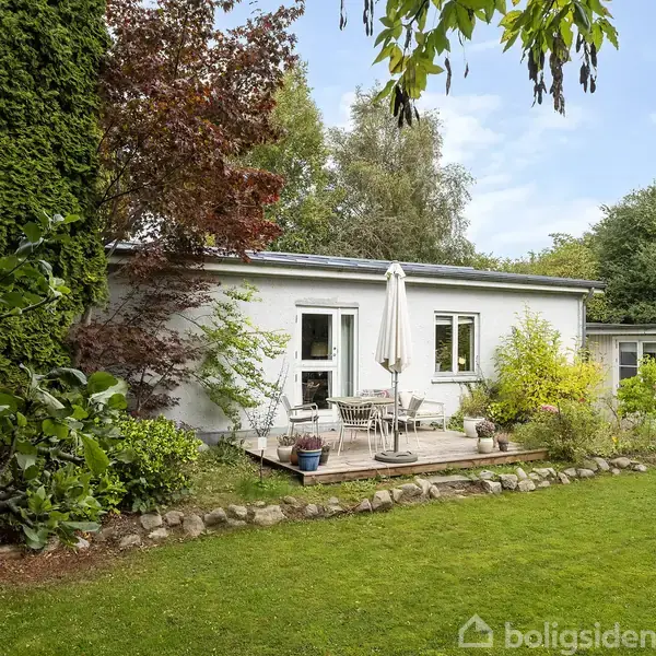 Et en-etagers hus har en lille terrasse med parasol og havemøbler. Græsplænen foran er omkranset af frodig vegetation og træer under en blå himmel.