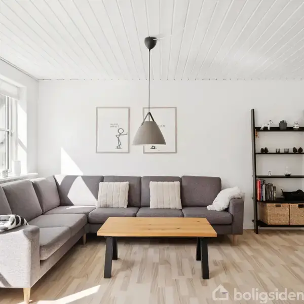 Gråt sofaarrangement står i hjørne af stue, belyst af naturligt lys fra vindue. Træbord foran, enkel sort hylde med dekorationer bagved. Minimalistiske kunstværker på væggen.