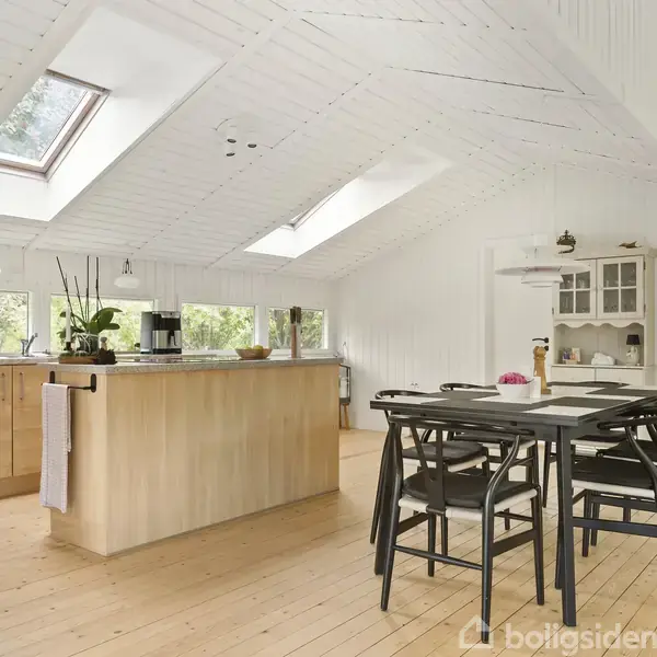 Køkkenø står i fokus med træoverflade, omgivet af spisebord og stole. Store ovenlysvinduer og planter giver et lyst og naturligt rum. Hvide skabe i baggrunden.