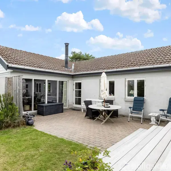 Huset har en terrasse med havemøbler og parasol. Terrassen rummer to liggestole. Det er omgivet af en græsplæne og har et solrigt miljø under en delvist skyet himmel.