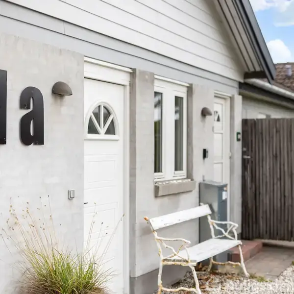 Husfacade med nummeret "1a" skrevet på væggen. En hvid bænk står foran huset, som har en lille hvid dør og to vinduer. Grusbelægning og planter omgiver huset.
