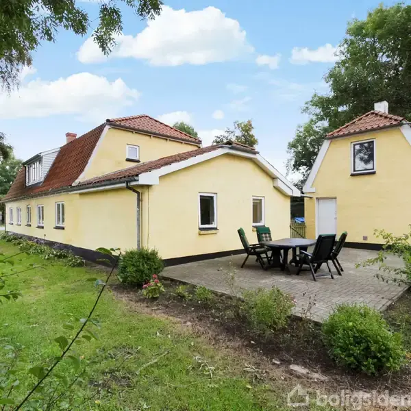 Et gulmalet hus med rødt tag står i en grøn have. En terrasse med bord og sorte stole ses ved siden af huset under en blå himmel med skyer.