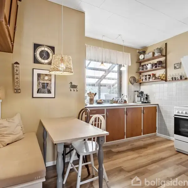 Et køkken med træbord og bænkesæde. Bordet er omgivet af to stole og en pude. Bagved er der køkkenskabe med vindue over. Væggen har dekorationer og et ur.