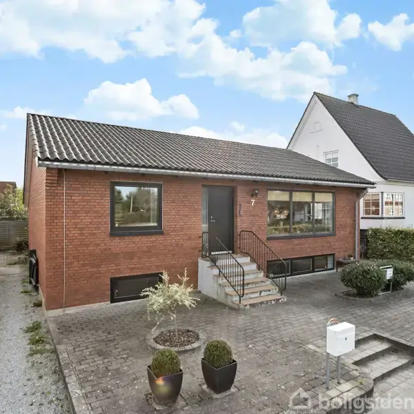 En murstensvilla med gråt tag står på en flisebelagt grund, flankeret af blomsterkrukker. Små trapper fører til hoveddøren. Naboen ses tæt ved, omgivet af hæk.