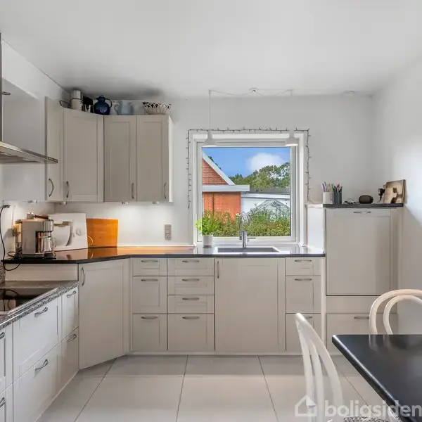 Et moderne køkken med hvide skabe og apparater, inklusive en ovn og emhætte. En pæn bordplade omringer en vask under et vindue med udsigt til en rød bygning udenfor.