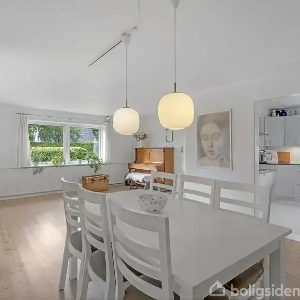 Et hvidt spisebord står i en spisestue, oplyst af to pendellamper. Bagved ses et klaver og et stort vindue med udsigt til en grøn have.