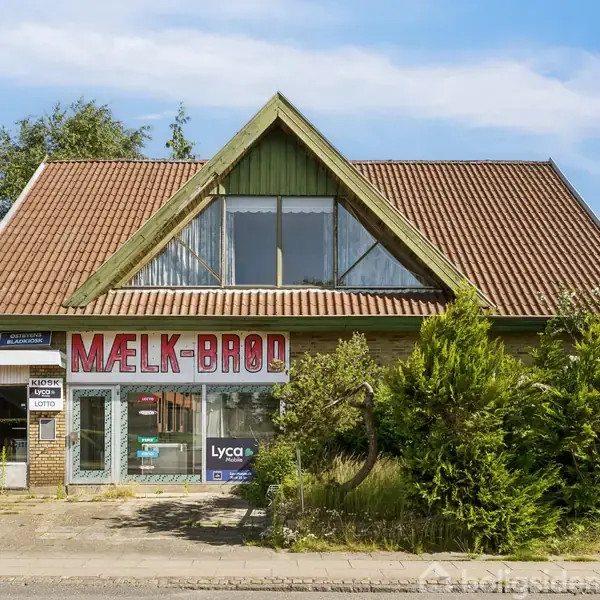 En butik med skiltet "MÆLK-BRØD" og "KIOSK" i en bygning med rødt tag. Nærliggende står grønne buske. Stedet ser ud til at være en lille lokal købmand.