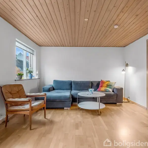 Sofa står stille i en lys stue med trægulve og træloft. Ved siden af er en lænestol, et rundt bord, en lampe og et vindue med planter.