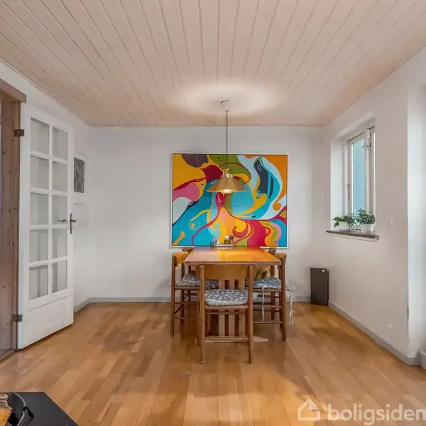 Et spisebord med fire stole står under en farverig, abstrakt maleri på væggen. Rummet har lyst trægulv, hvidmalede vægge, og et stort vindue til højre.