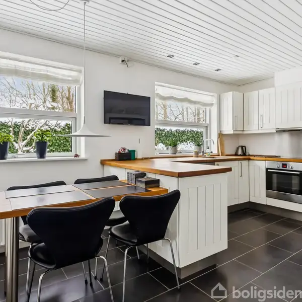 Et moderne køkken med hvide skabe. En spiseplads med sorte stole ved træbord. Vinduer viser en grøn hæk udenfor. Sort flisegulv og hvidt loft.