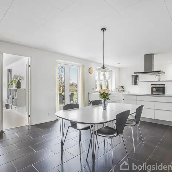 Rundt spisebord med sorte stole står i moderne køkkenalrum. Et skab, komfur og ovn ses bagved, mens store vinduer og glasdøre giver udsigt til havebelysning. Ingen synlig tekst.
