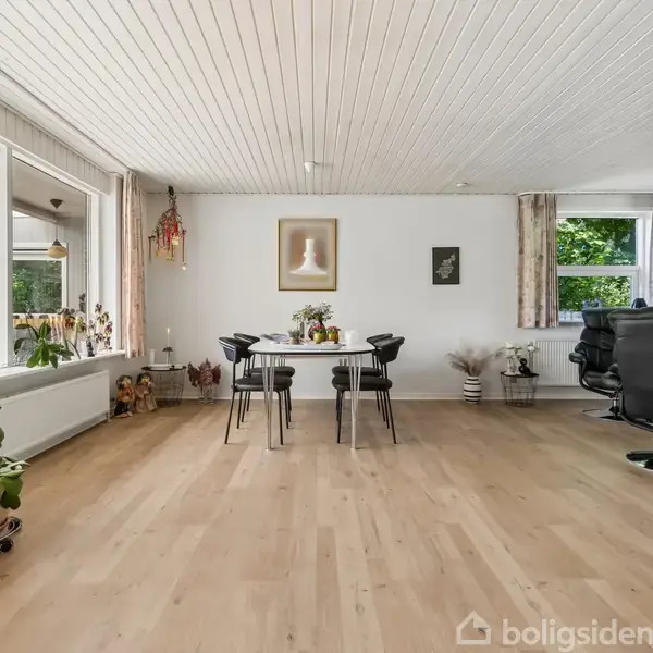 Spisestue med hvidt loft og trægulv, hvor et rundt bord med sorte stole står midt i rummet. Store vinduer giver udsigt til udestue; planter pynter rummet.