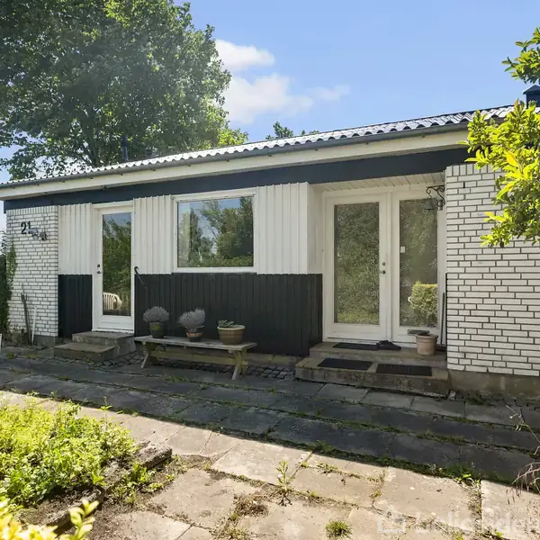 Hus med hvid murstensfacade og tre store vinduer, omgivet af grønne træer og buske, står på en flisebelagt terrasse. Himlen er blå med spredte skyer.