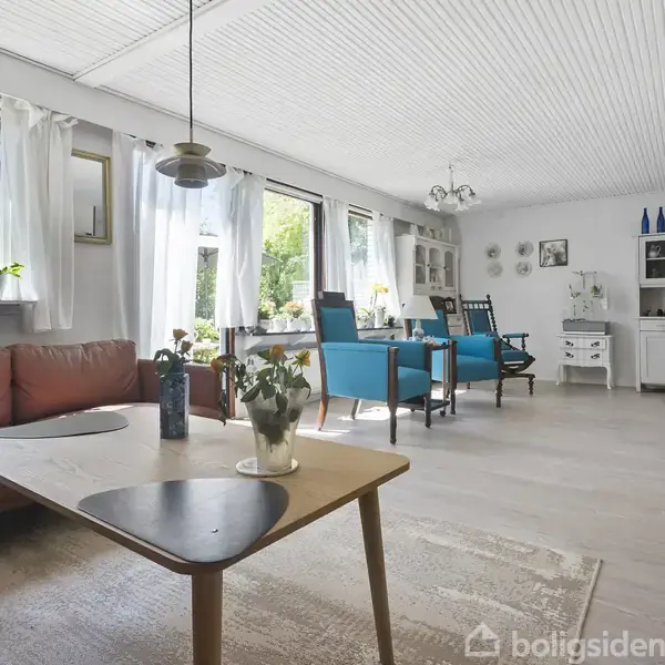 En lys stue med sofaer i blå og rød ved et træbord. Vinduer med gardiner slipper lys ind, og planter står i vindueskarmen. Møbler i klassisk stil fylder rummet.