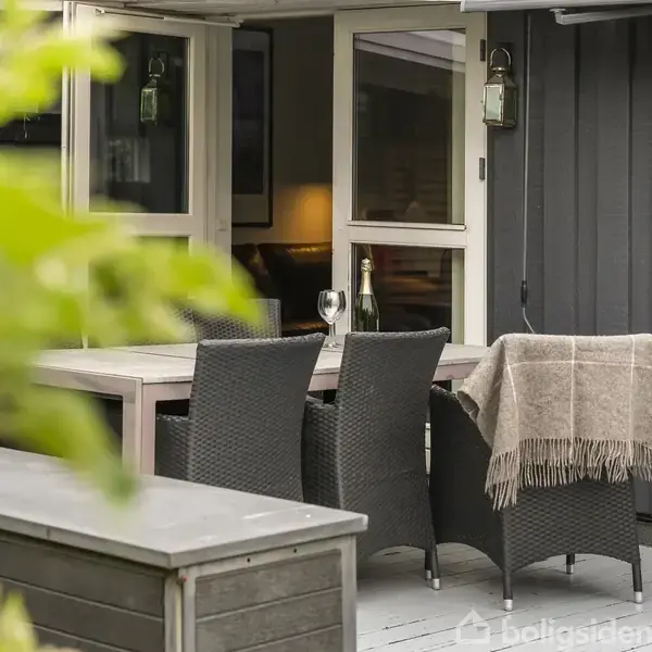 Et udendørs terrasseområde med et bord og fire fletstole, hvoraf én er dækket med et plaid. Der er et glas og en flaske på bordet. Åbne døre viser indendørs belysning.