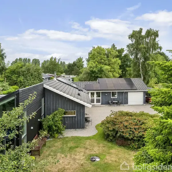 Et sort træhus med solpaneler på taget står omgivet af frodige grønne træer og buske. Foran huset er en grusbelagt gårdsplads. Himmel er let skyet.