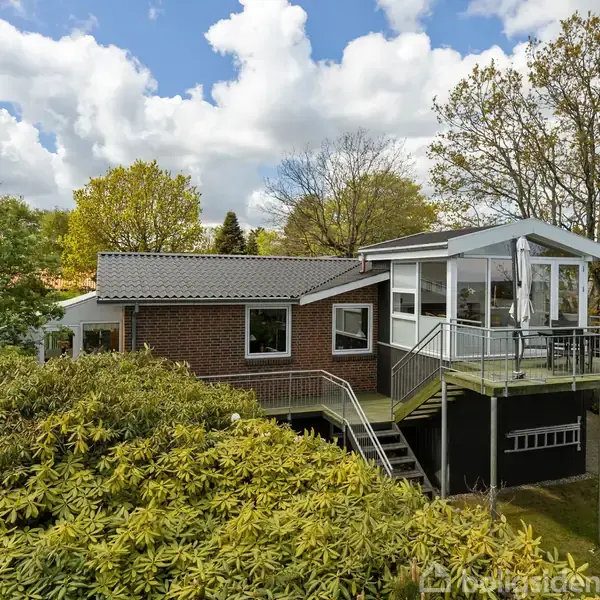 Huset er en moderne murstensvilla med store vinduer og en terrasse. Det ligger omgivet af frodig vegetation og træer, under en skyfri himmel med få skyer.
