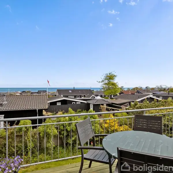 En terrasse med et rundt bord og fire stole står tom i solskin. Baggrunden viser en række tagtoppe og en udsigt over havet under en klar blå himmel.