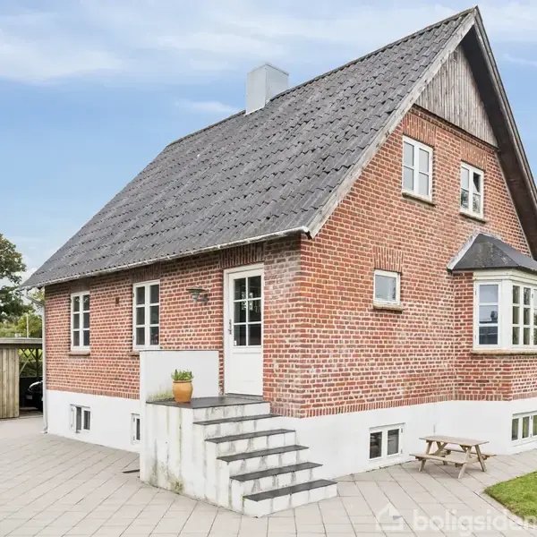 En rød murstensvilla med gråt sadeltag har en trappe op til hoveddøren. I forgrunden er en flisebelagt terrasse, og omgivende græsplæne skaber en hyggelig haveatmosfære.