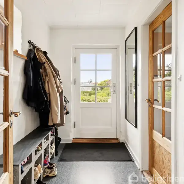 Gangen har en bænk med skohylder og jakker på væggen. Der ses en hvid dør med glasruder mod en grøn udsigt. Rummet er lyst og velorganiseret.