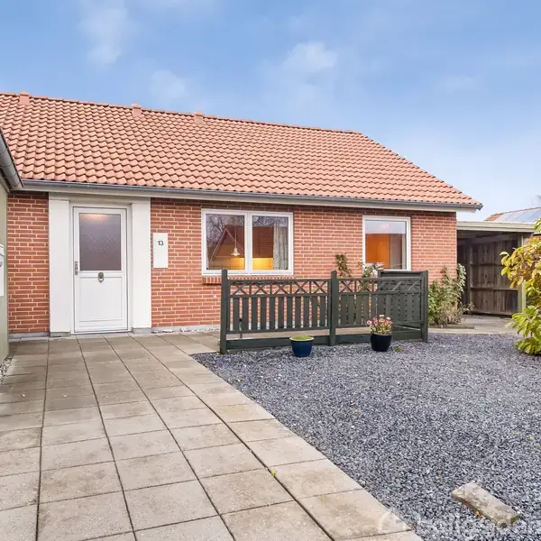 Huset er en lav, rød murstensbygning med orange tegltag. Det står ved siden af en flisebelagt indgang og en grusbelagt have med planter.