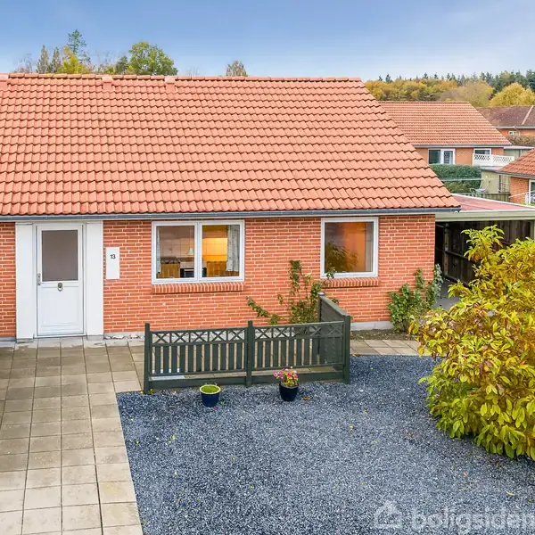 Rødt murstenshus med rødt tegltag står på en velholdt grund. Foran huset er en flisebelagt sti, grusbelægning og blomsterbed. Området er beboelsesområde med andre huse omkring.