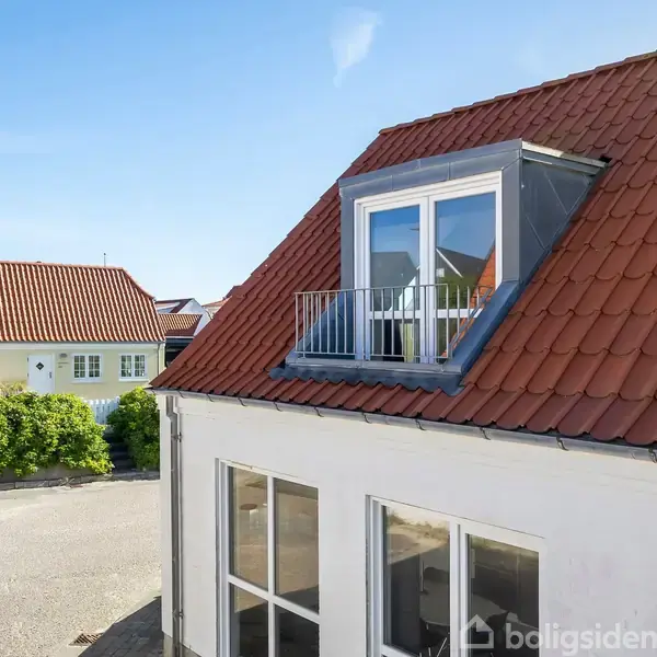 Rødt tegltagshus med kvist og fransk altan i et beboelsesområde. Solskin oplyser facaden, mens omkringliggende huse og grønne buske bidrager til nabolagets atmosfære.