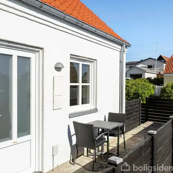 Et lille, hvidt hus med rødtag står i sollys. En havegård med sorte plankeværker og et lille bord med to stole står op ad husmuren.