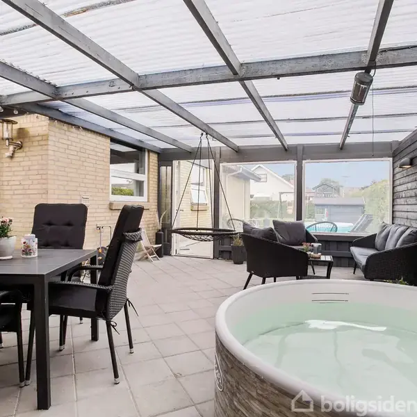 Et overdækket terrasseområde indeholder et spisebord med fire stole, en moderne jacuzzi, og en sofagruppe. Lyse murstensvægge og glaspartier skaber en lys og afslappet atmosfære.