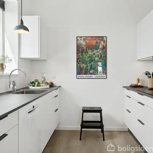 Et moderne køkken med hvide skabe og sorte bordplader. En sort skammel står ved en væg, der har en farverig plakat med teksten "SUSANNE NØRGAARD" "LOUISIANA". Sollys strømmer ind gennem vinduet.