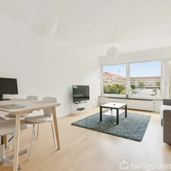 En lys stue med trægulv, et sofabord på et tæppe, en sofa ved det store vindue, en tv-skærm i hjørnet og et skrivebord med computer.