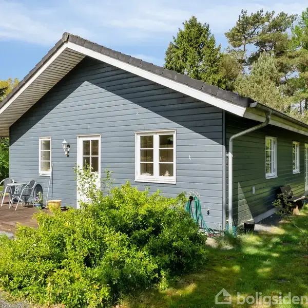 Gråt hus står stille omgivet af frodig grøn have og træer. Foran huset ses en lille terrasse med et par stole og planter.